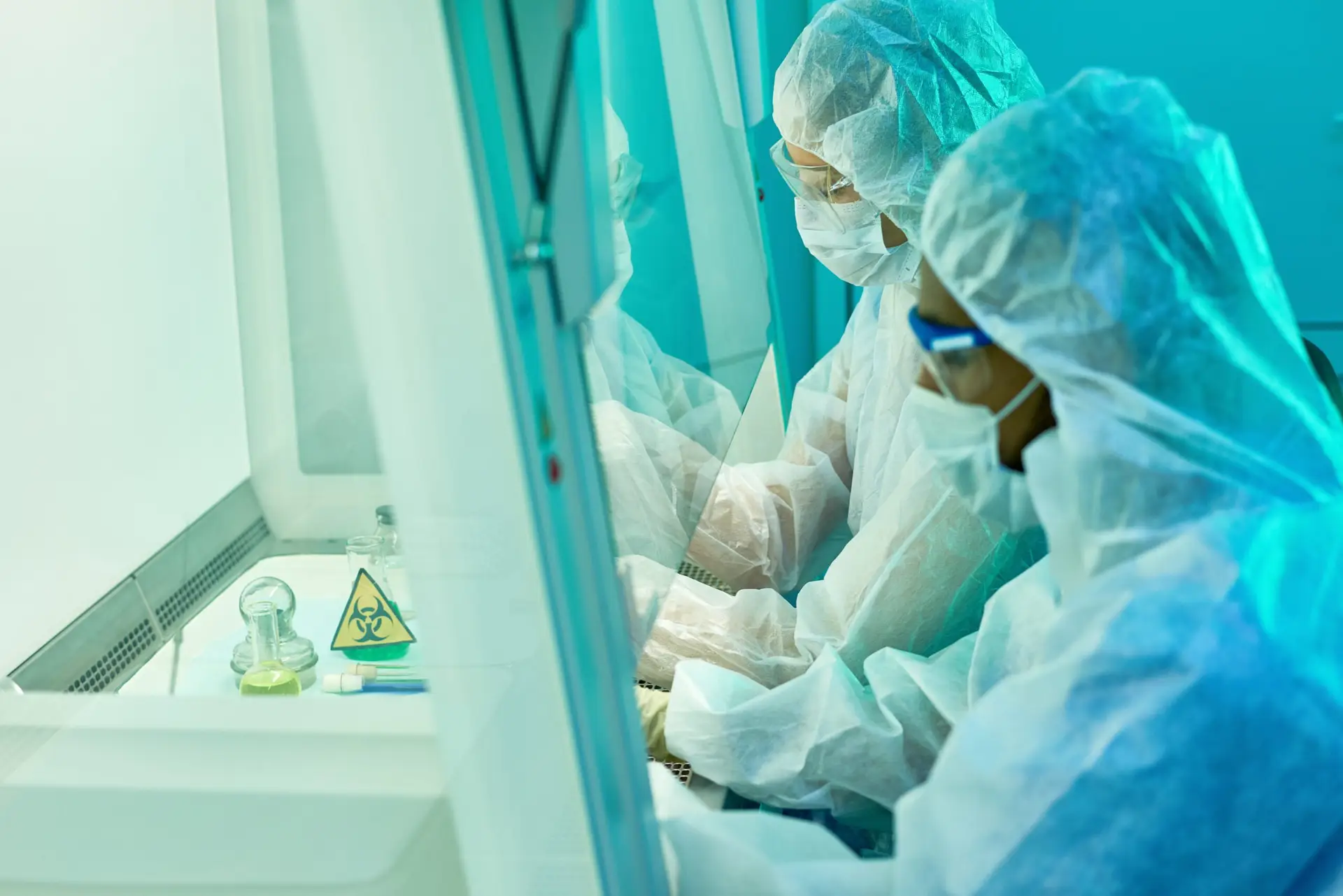 Concentrated unrecognizable laboratory colleagues sitting by special desk with transparent glass and holding dangerous chemical experiment